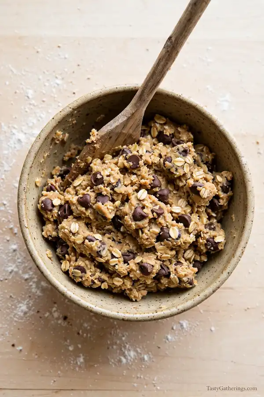 Top view of oatmeal chocolate chip cookie dough in a rustic bowl with oats and chocolate chips, wooden spatula on light wooden table.