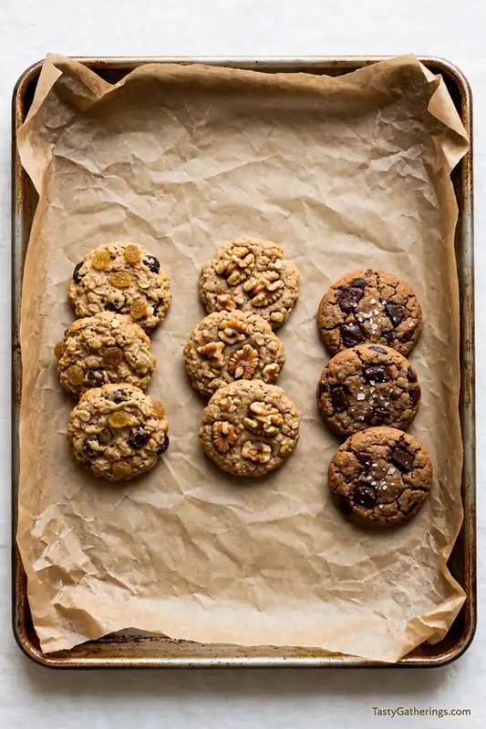 Baking tray with three variations of oatmeal cookies - raisin, walnut, and dark chocolate sea salt on parchment paper.